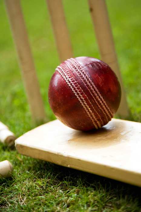 Red leather cricket ball on grass with stumps.