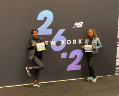 Marie Young (left) and Marva Joseph, standing in front of a sign that has 26.2 written on it. They are holding their participant stickers.