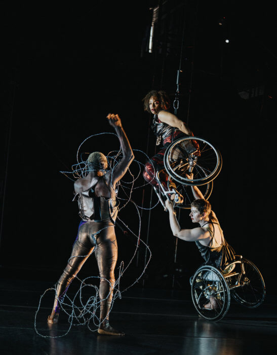 Three dancers face each other onstage. Jerron Herman, a Black man with blonde hair, stands boldly facing the others, his body tense with energy. His tight pants and leather top shimmer. His fist flies overhead as silver barbed wire cascades from head to feet. Alice Sheppard and Laurel Lawson are stacked and lean in toward Jerron with concentrated expressions. Alice, a multiracial Black woman with coffee-colored skin and short curly hair, hovers in the air. Laurel, a white dancer with cropped hair, balances beneath her; she grips Alice’s wheels while tilting on one wheel. 