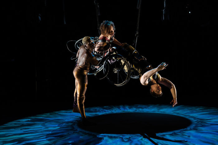 Three dancers are encircled by waves of green and blue light, designed by Michael Maag. Laurel Lawson, a white dancer with cropped hair, bends backward, arms curved over head and torso, as she hovers above the stage. Alice Sheppard, a multiracial Black woman with short curly hair and coffee-colored skin, also hovers behind Laurel, partnering her and cradling her legs and wheels. Jerron Herman, a Black man with blonde hair, balances on one foot, embracing Alice and Laurel’s wheels and peering over at Laurel with curiosity. 