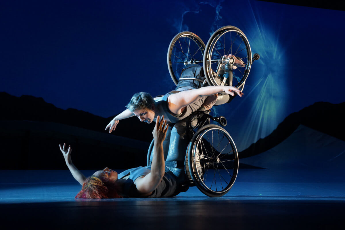 Laurel Lawson, a white dancer with short cropped teal hair, is balancing with arms spread wide, wheels spinning, and supported by Alice Sheppard. Alice, a multiracial Black woman with coffee-colored skin and curly hair, is lifting from the ground below. They are making eye contact and smiling. A burst of white light appears in a dark blue sky.
