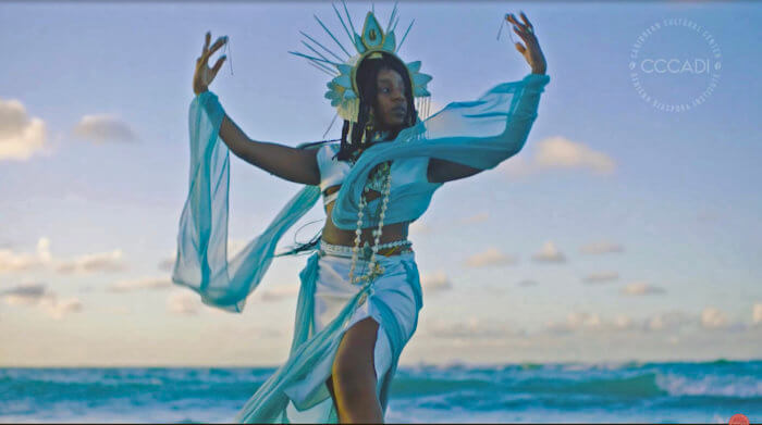 Screen grab of a scene in Alexis Garcia's film "Daughter of the Sea" (2022). This shows the spiritual goddess of the film, Yemaya, who is wearing all blue attire, standing with her back towards the ocean.