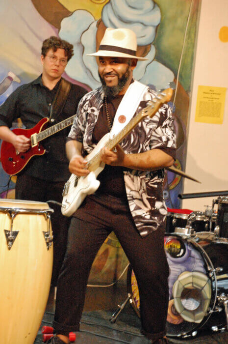 Nkumu Katalay, on bass guitar, wearing a cream hat. A member of his Life Long Project band is standing behind him on stage. 