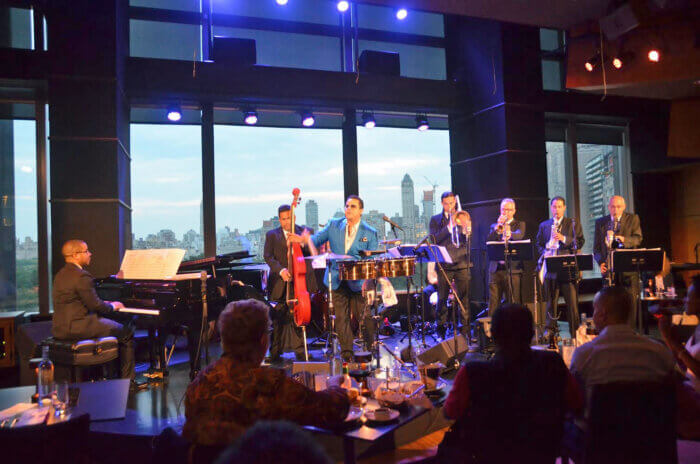 Tito Puente Jr. and Willy Rodriguez are on stage performing with a band during an event at Lincoln Center in May of this year. Rodriguez is playing the piano, and Puente Jr. is singing. 