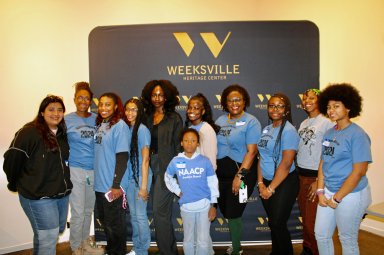 A diverse group of students stand against the Weedsville Heritage Center sinage during a "Call to Action” expo to commemorate the late iconic leader Dr. Martin Luther King Jr.'s lasting legacy. Deputy Director Sasha Miller, second from left, and chief of Programs of The Legacy Network, Jennella Young, seventh from left, are pictured with the students.