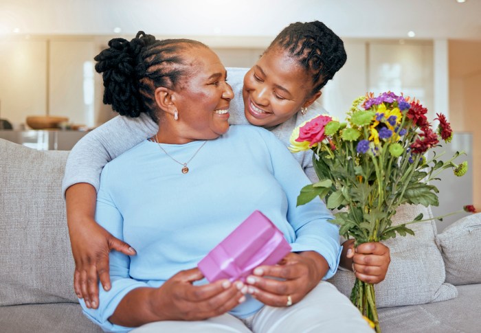 Black woman, mothers day and hug with flowers on sofa for love, box or gift to mom in home. Woman, bouquet and senior mama with wow, smile or happy for surprise, celebration or birthday on couch