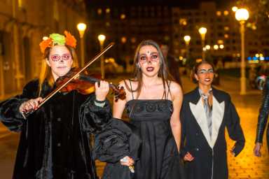A group of people dressed in Halloween costumes walking down a street with one of them playing a violin.
