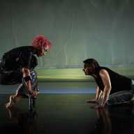 Alice and Tati face off. Alice is balancing on her short crutches, legs bent and folded under herself, tops of her toes grazing the floor. They peer at each other with curiosity and intensity.