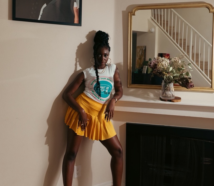 A photo of wearing heels, standing up against a wall with her right knee bent, and her right arm hanging off of a ledge.