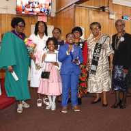 Valcia Williams's son, Nicholas Charles, holds a plaque, and daughter, Noelani Charles, holds a certificate. Valcia Williams is in a white usher dress; her mother, Catherine Williams, is left. They are flanked by members of the UWF Special Recognition Committee. UWF President Marlene Ferguson is at right.