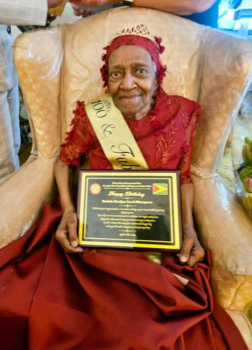 One-hundred-year-old Eulah Evelyn Levi-Marques celebrated with Flowers, a fruit-basket, a plaque, and Golden Arrowhead flag Pin from Guyana Consul General Ambassador Michael E. Brotherson. She was decked out in a fancy dress to celebrate her milestone birthday on April 28, in Brooklyn. Here she is with a beautiful smile, while holding a Plaque from Guyana Consul General to New York Ambassador Michael E. Brothreson.